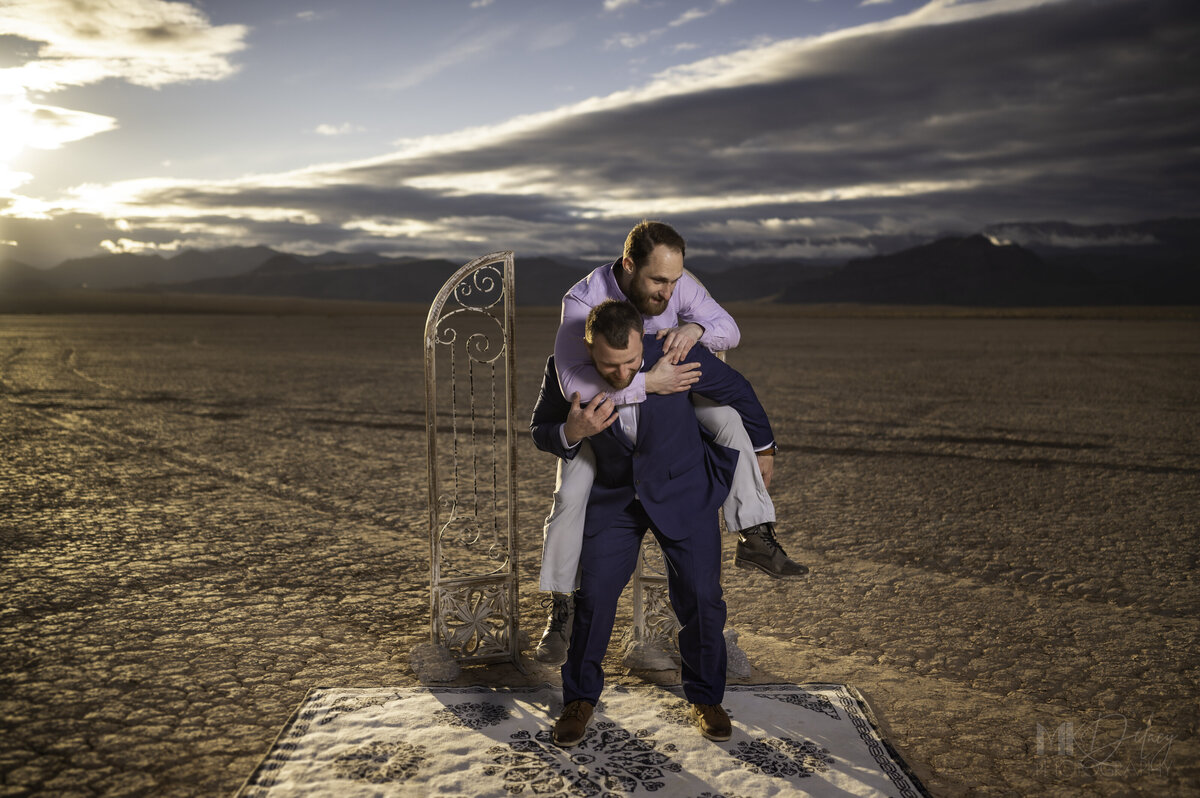Groom and best man silly jumping on grooms back elopement pictures Dry lake Bed elopement Blue Suit on Groom  flowers by michelle  bride in cream color wedding dress with deep  plunging  neckline mountain skyline  sunset las vegas wedding photographers mk delacy photography