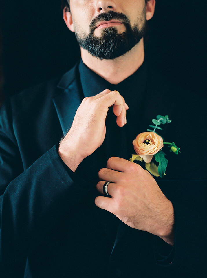Close-up of groom wearing a black tuxedo with ivory flower adjusting his tie.