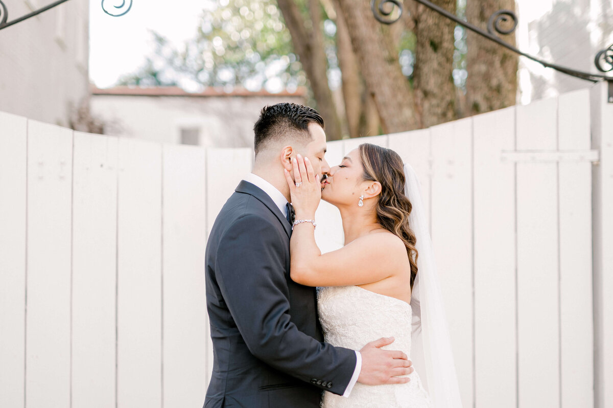 Bride and groom kissing in front of white fence during wedding portraits