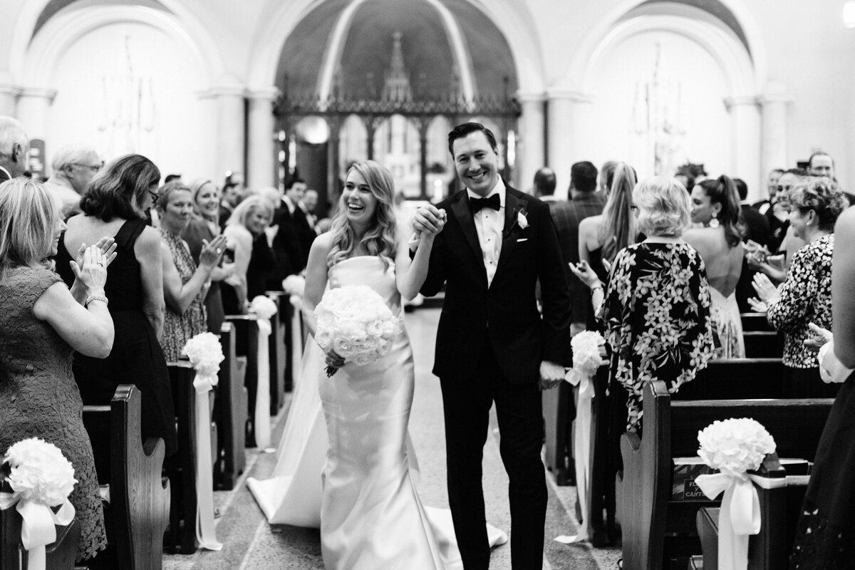 Bride & groom holding hands & walking down aisle in Catholic church recessional surrounded by congratulatory guests