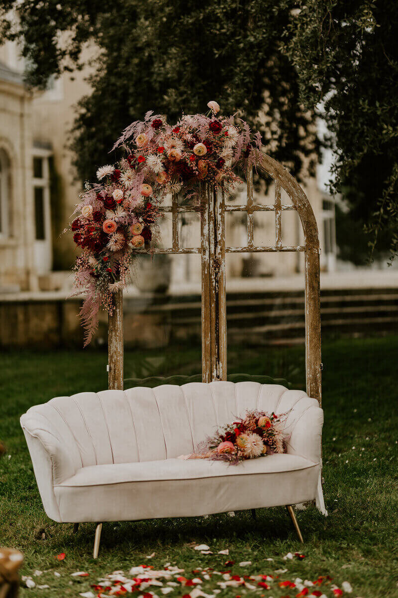 Bouquet de fleur posé sur un canapé blanc vintage. Décor verdoyant en arrière plan et arche en bois surmontée d'une composition florale. Photo prise par Laura, photographe professionnelle.