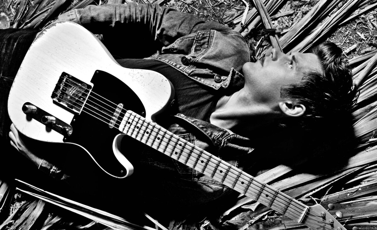 Male musician portrait black and white Dave Green wearing jean jacket holding white electric guitar while lying down against palm branches