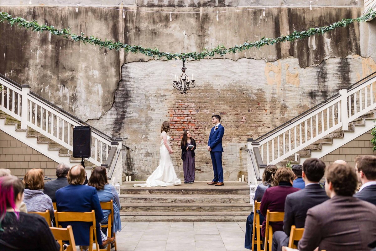A bride and groom stand with an officiant on a small stage between two staircases, exchanging vows outdoors. Guests are seated, watching. The backdrop is a weathered brick wall with garland and a chandelier overhead.