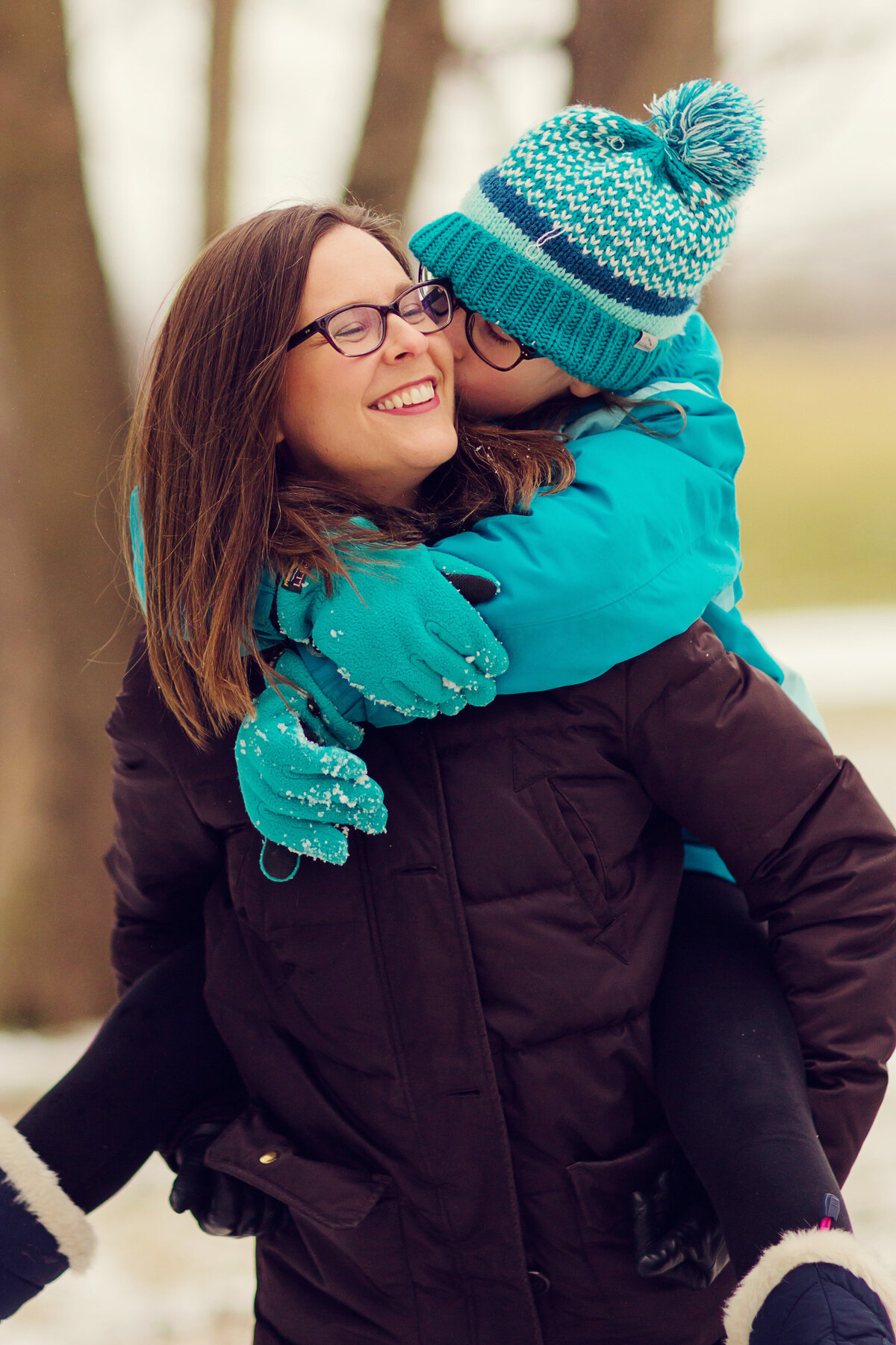 Mom in a black coat and glasses has her daughter on her back. Her daughter is wearing a blue coat and blue hat.