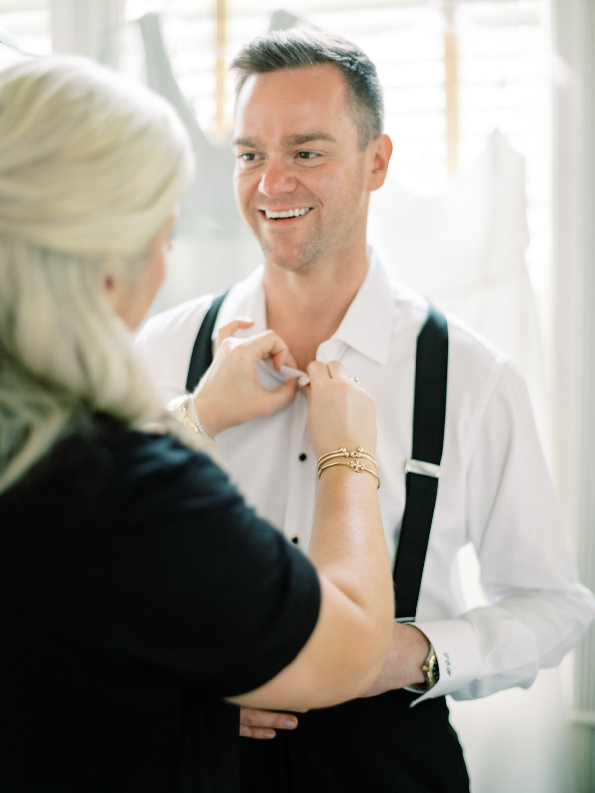 A joyful groom is assisted with his outfit by a caring individual, capturing a special moment of preparation before a wedding ceremony filled with excitement.
