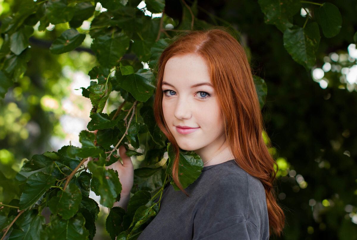A close up portrait of a beautiful blue eyed redhead teen girl wearing a dark gray shirt is surrounded by greenery. Photograph by Dynae Levingston.