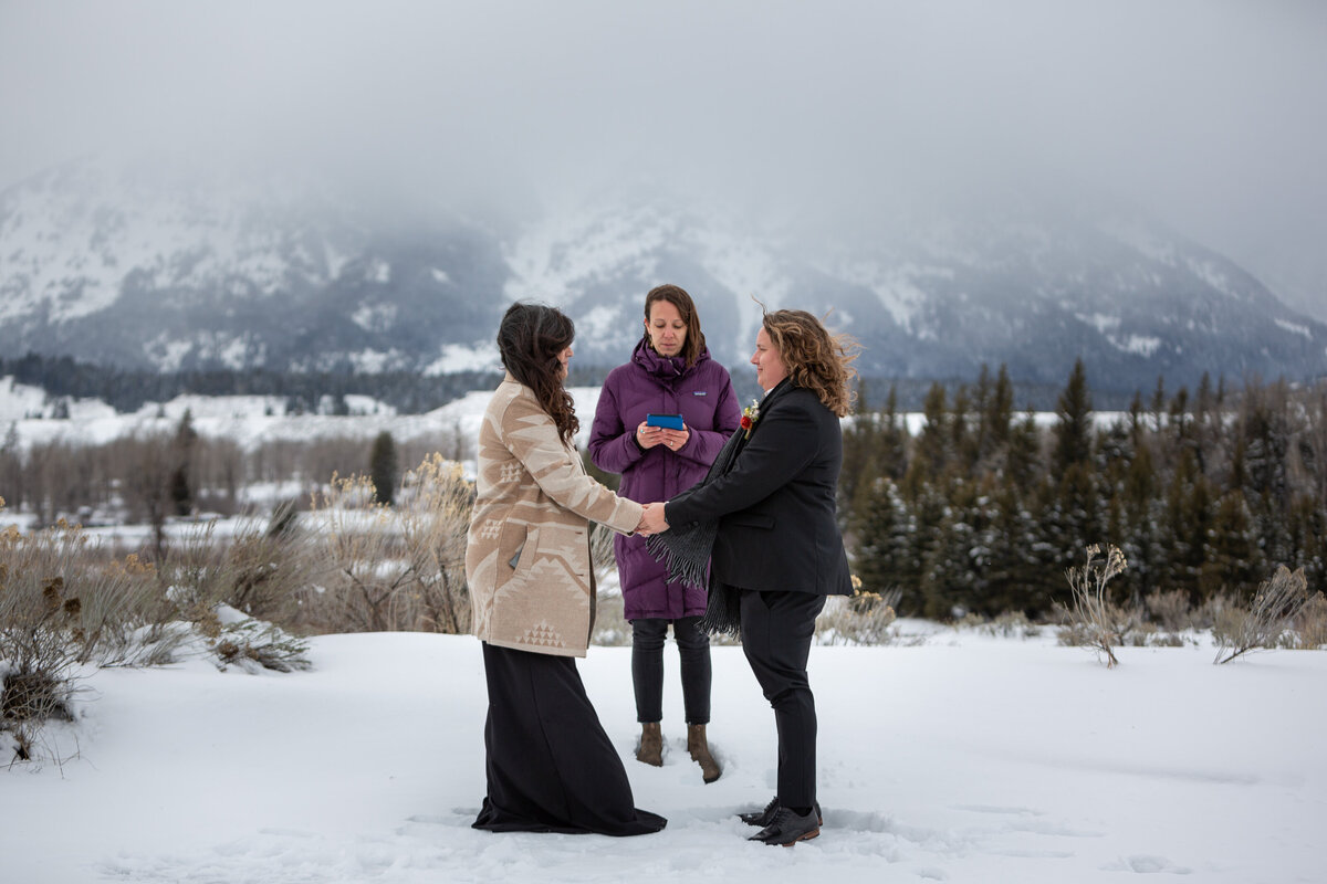 Two brides stand facing each other and holding hands while their officiant reads behind them on their snowy elopement day in Grand Teton National Park.