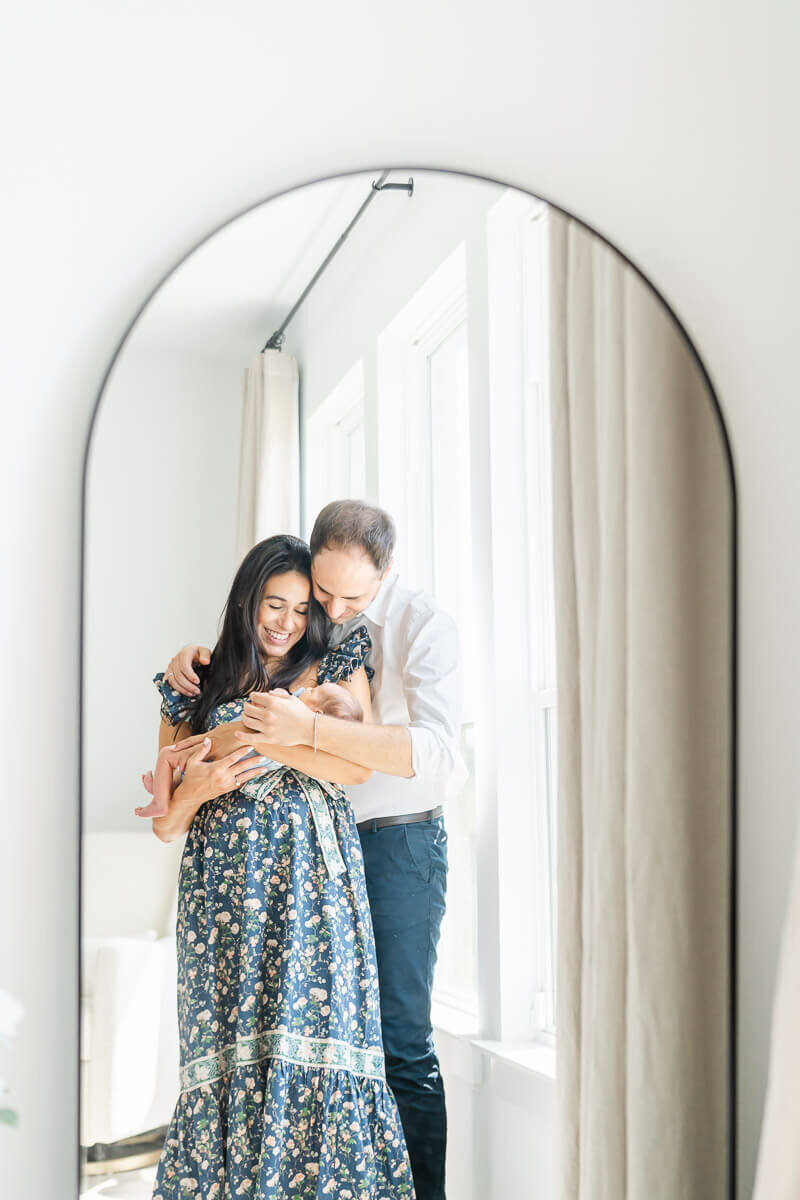 a couple stands in front of a mirror and look down at their newborn.