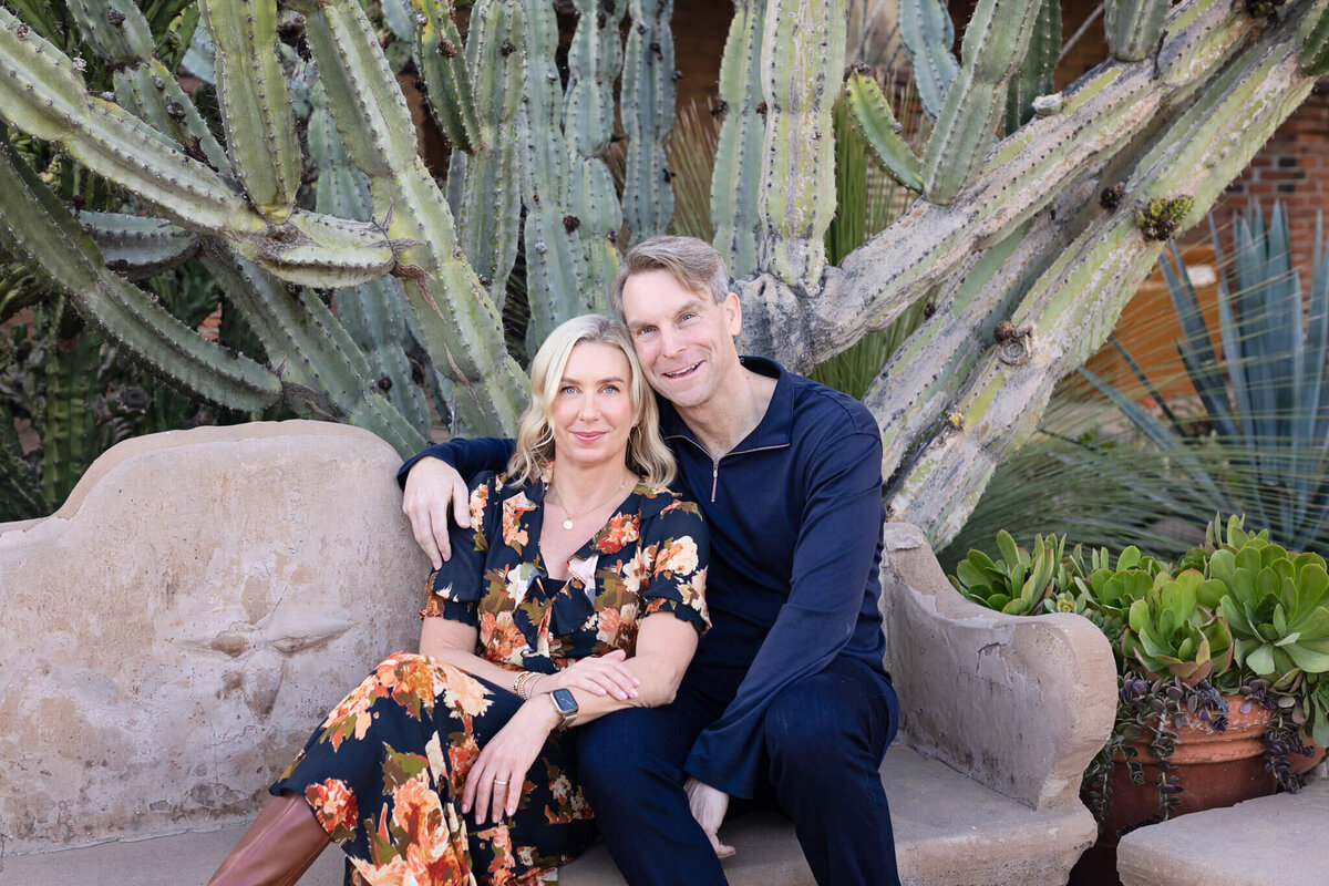 A woman with blonde hair wearing a navy and orange floral dress with her back leaning against her husband's chest who's wearing a navy long sleeve shirt with his arm around his wife, smiling at the camera while they sit on a park bench.