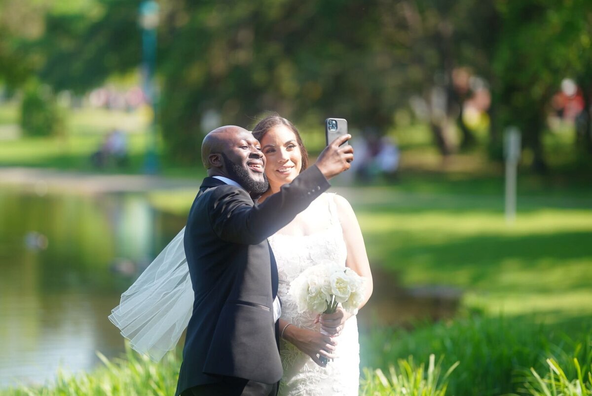 A couple smiles and takes a selfie in a picturesque nature park. The lush green surroundings and their joyful expressions capture a moment of fun and connection amidst the natural beauty.