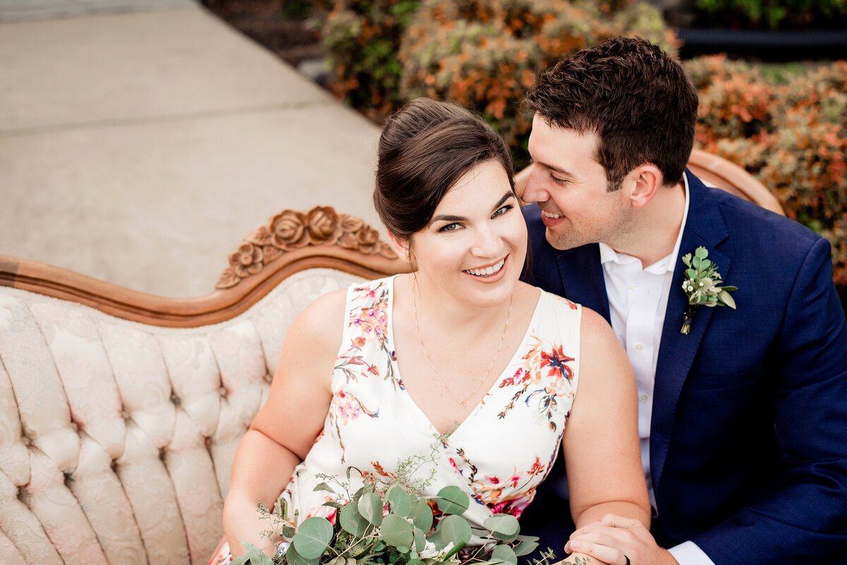 Bride wearing a floral dress and smiling at the camera while her new husband whispers in her ear, they are both sitting on a vintage couch