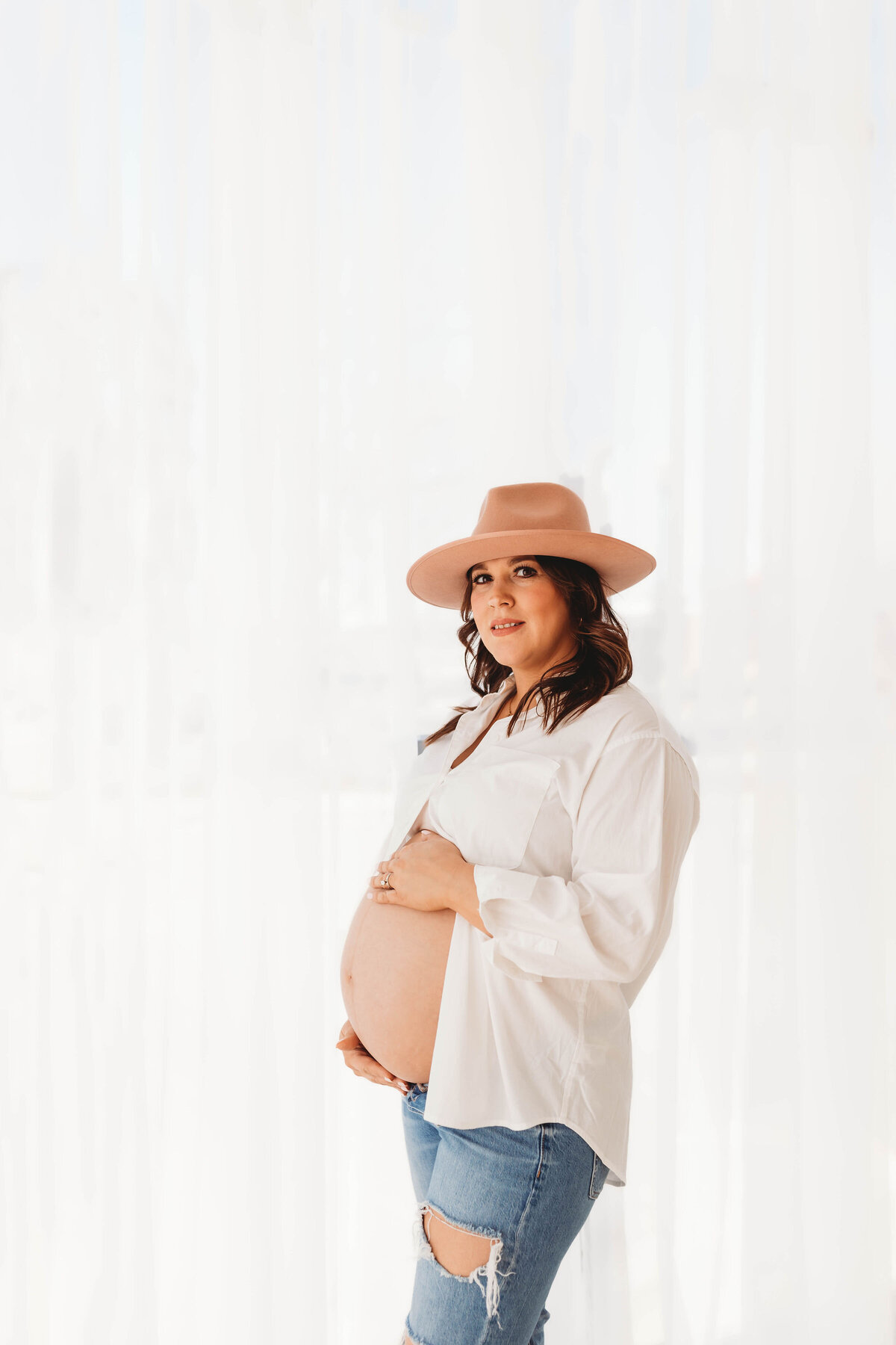 Pregnant mom in a white button up shirt with jeans and a hat holds her bare belly.