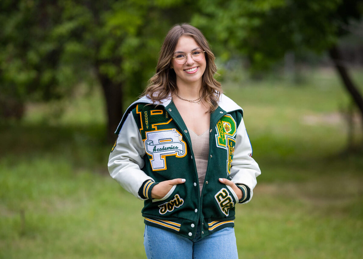 high school senior girl wearing her letterman jacket standing near trees