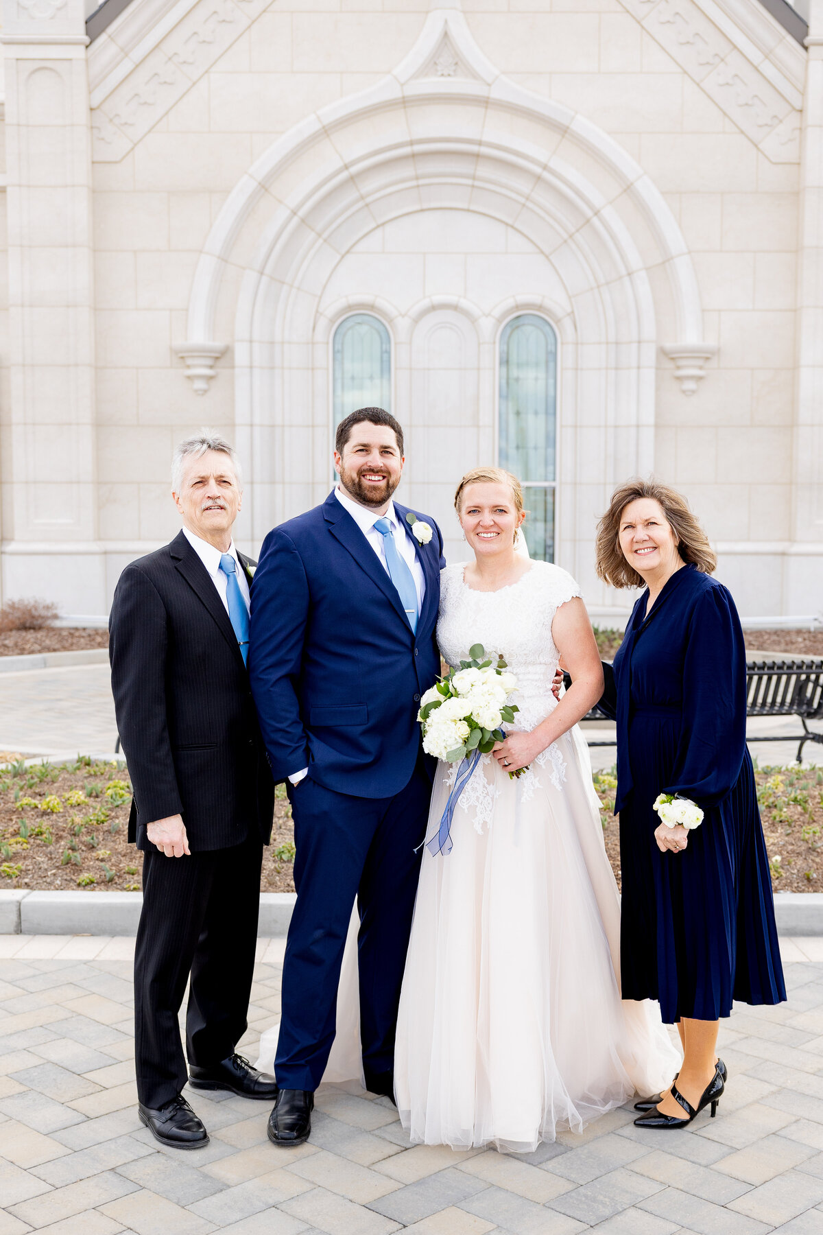 Bride and groom pose together with the groom’s parents, all dressed formally and smiling in front of the temple’s arched windows.