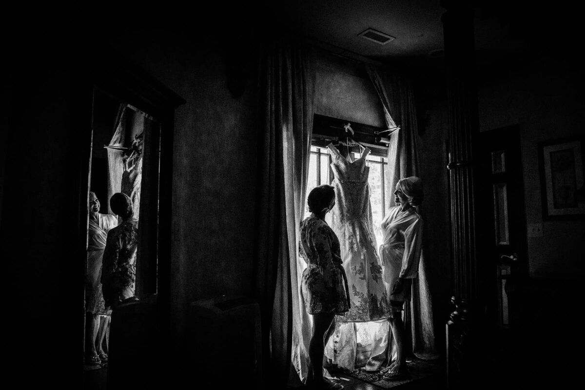 A dramatic black and white image of a wedding dress hanging in a window, with the bride and bridesmaids preparing in the background.