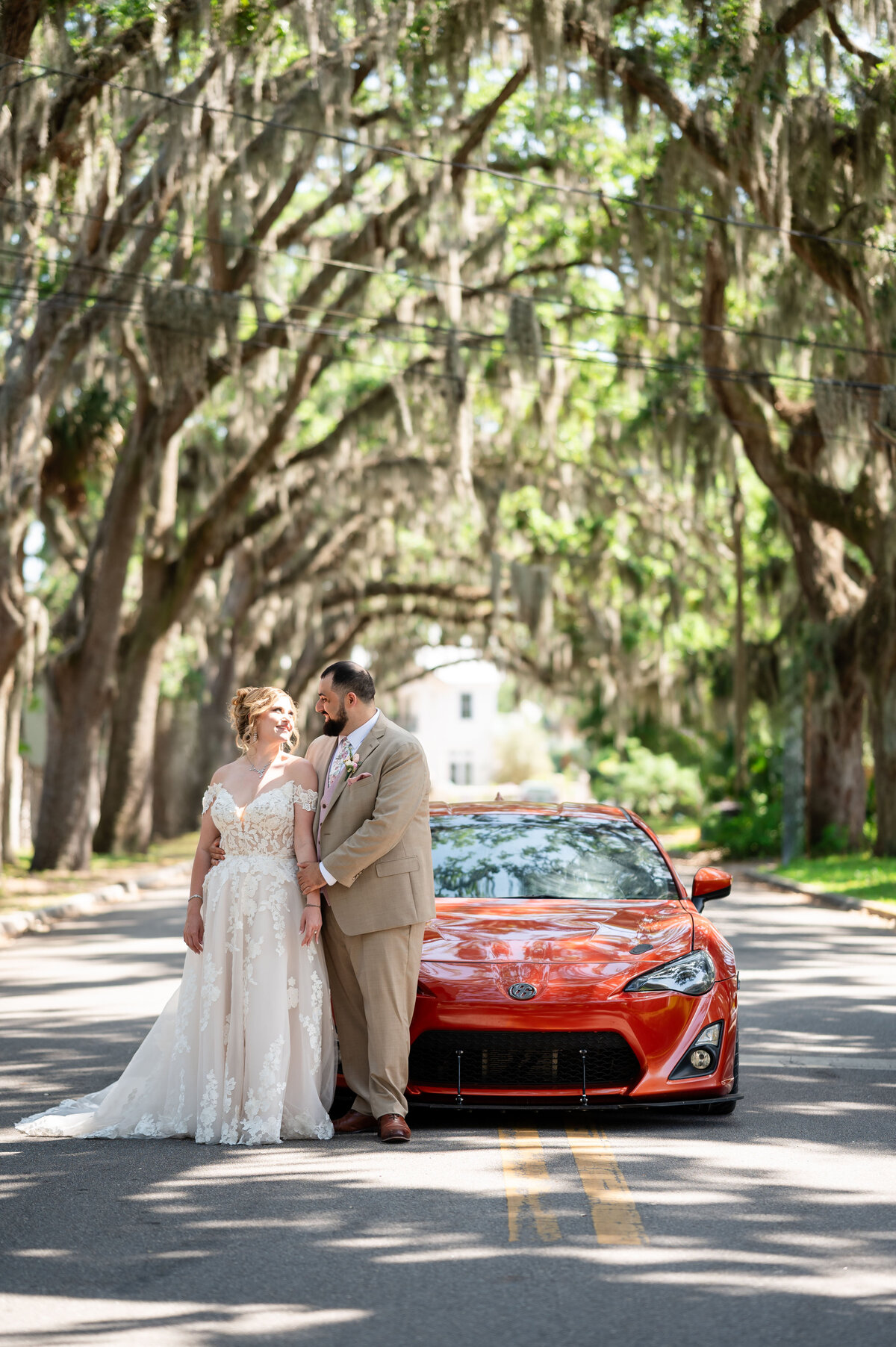 A bride and groom share a quiet moment in front of a vibrant orange sports car on the iconic Magnolia Avenue in St. Augustine, Florida, just outside the historic Fountain of Youth. Surrounded by a tunnel of moss-draped oak trees, the couple stands closely together—she in an off-the-shoulder lace gown, and he in a tan suit. This romantic, tree-lined backdrop was beautifully captured by Heartful Films, a Jacksonville wedding photographer and videographer specializing in timeless, location-rich wedding storytelling.