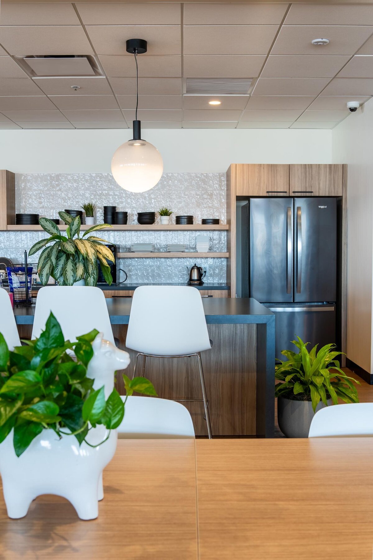 Modern office kitchenette with wood cabinetry, open shelving, black refrigerator, and tabletop plants, with white chairs in the foreground.