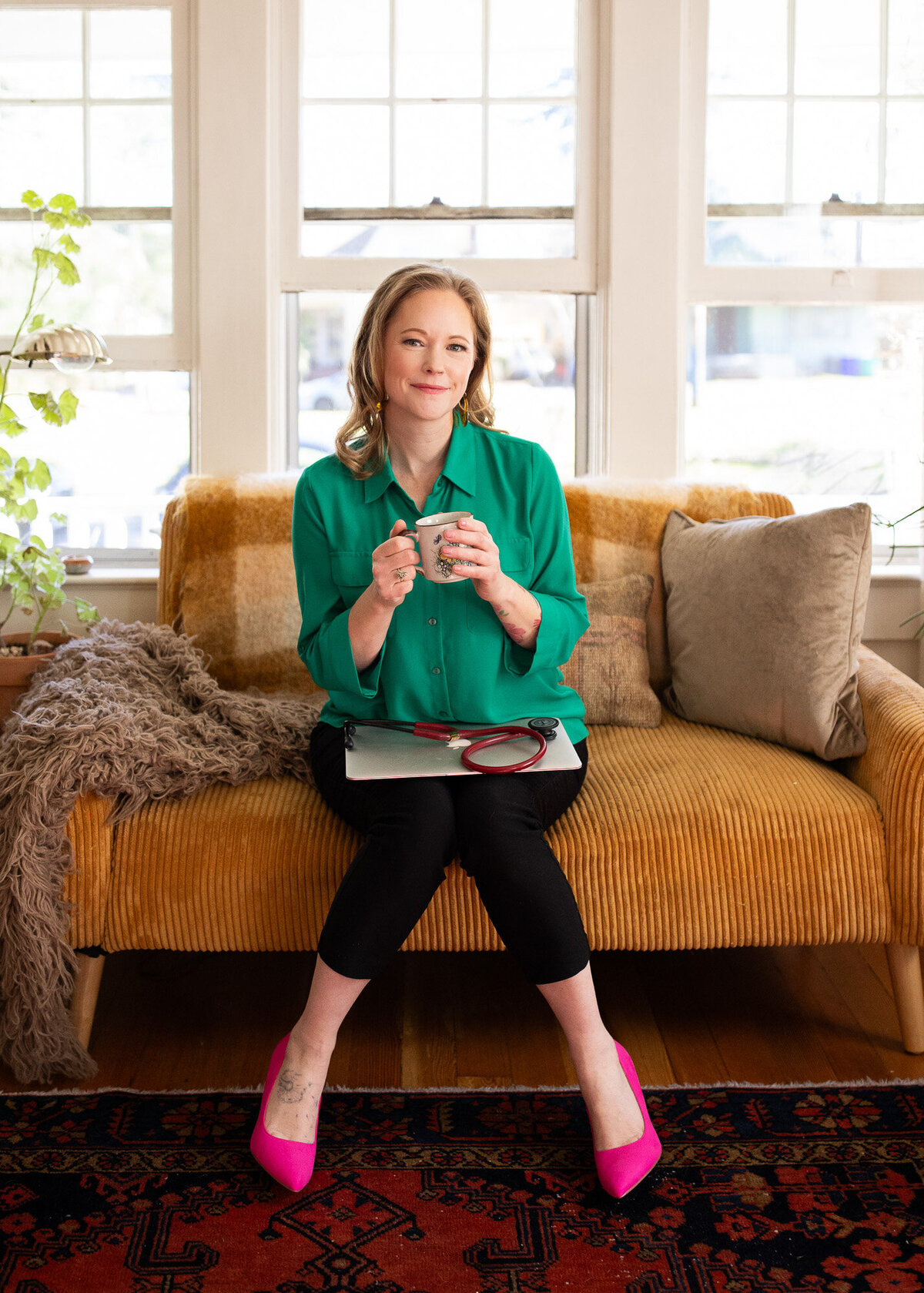 Woman holds coffee while she sits on couch and her laptop with stethoscope is on top resting. 