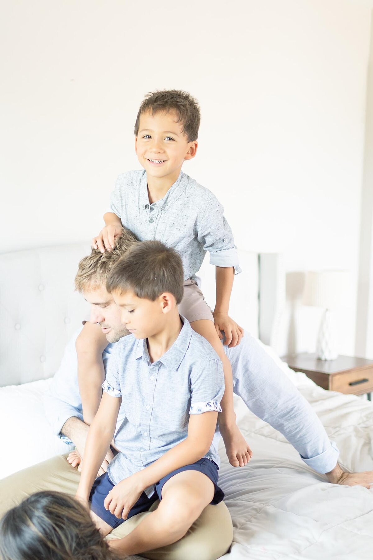 Photograph of a Dad sitting on the bed with his 2 sons, in blue short sleev button downs,  on sits on Dad's shoulders and the other sits on his lap by Family Photographer in Virginia Beach Mary Eleanor