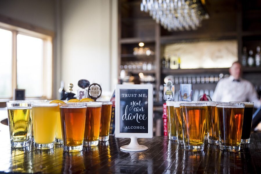 A bar with several glasses of beer of various shades of amber with a small chalkboard easel sign that says "Trust me, you can dance. - Alcohol"