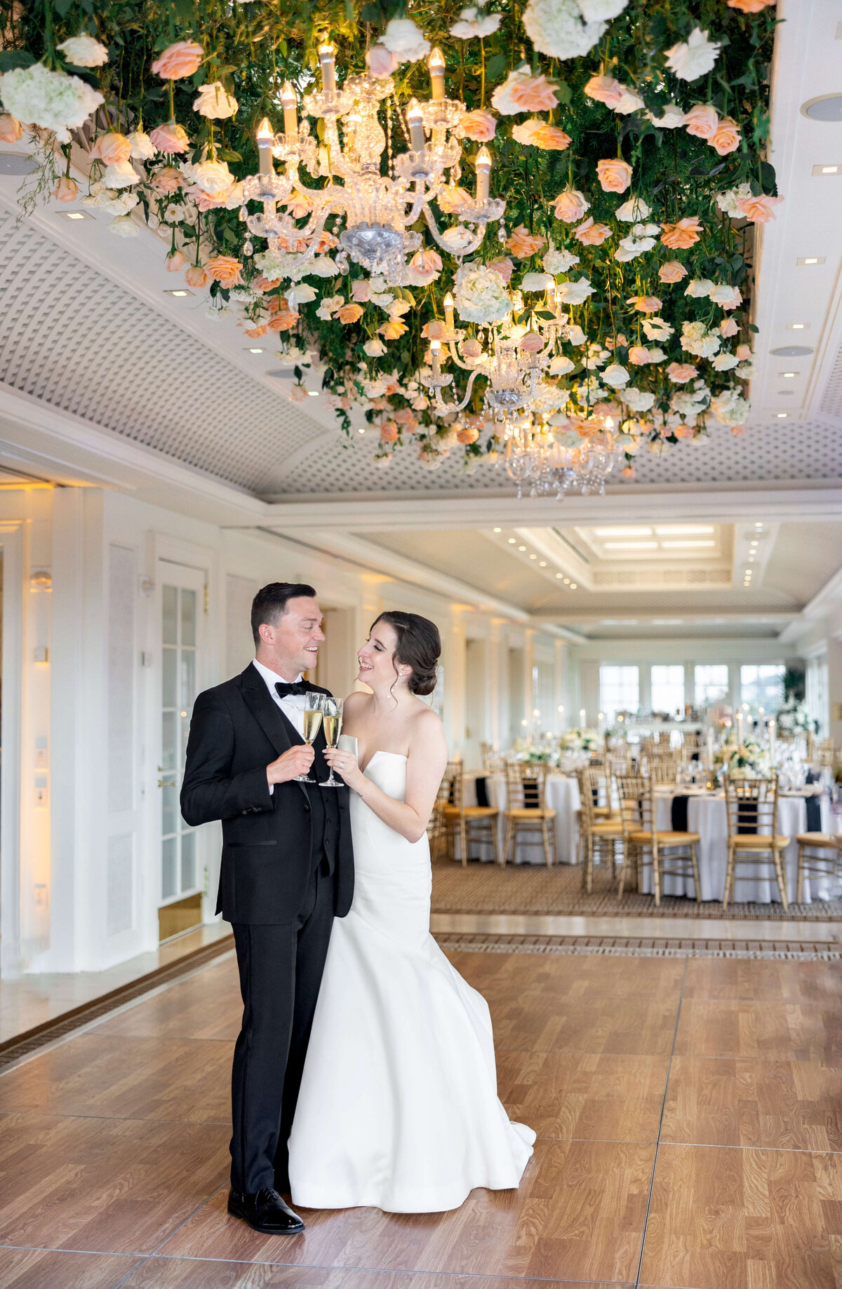 A bride and groom stand on a wooden floor under chandeliers adorned with flowers. The bride wears a white gown, and the groom wears a black suit. Theyre clinking champagne glasses, with elegantly set tables in the background.