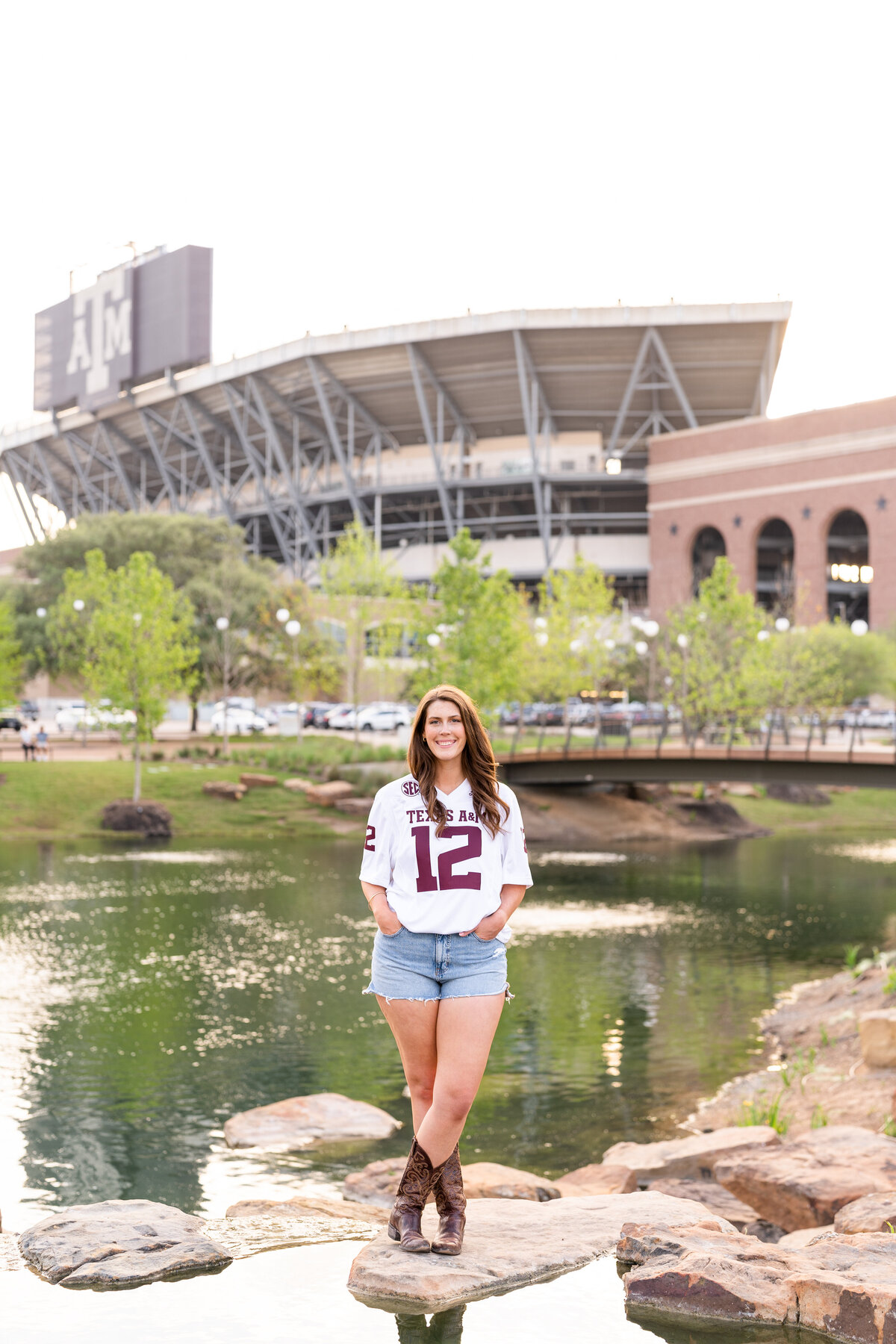 Texas A&M senior girl smiling with hands in pocket and standing on rocks in Aggie Park while wearing white jersey in front of Kyle Field