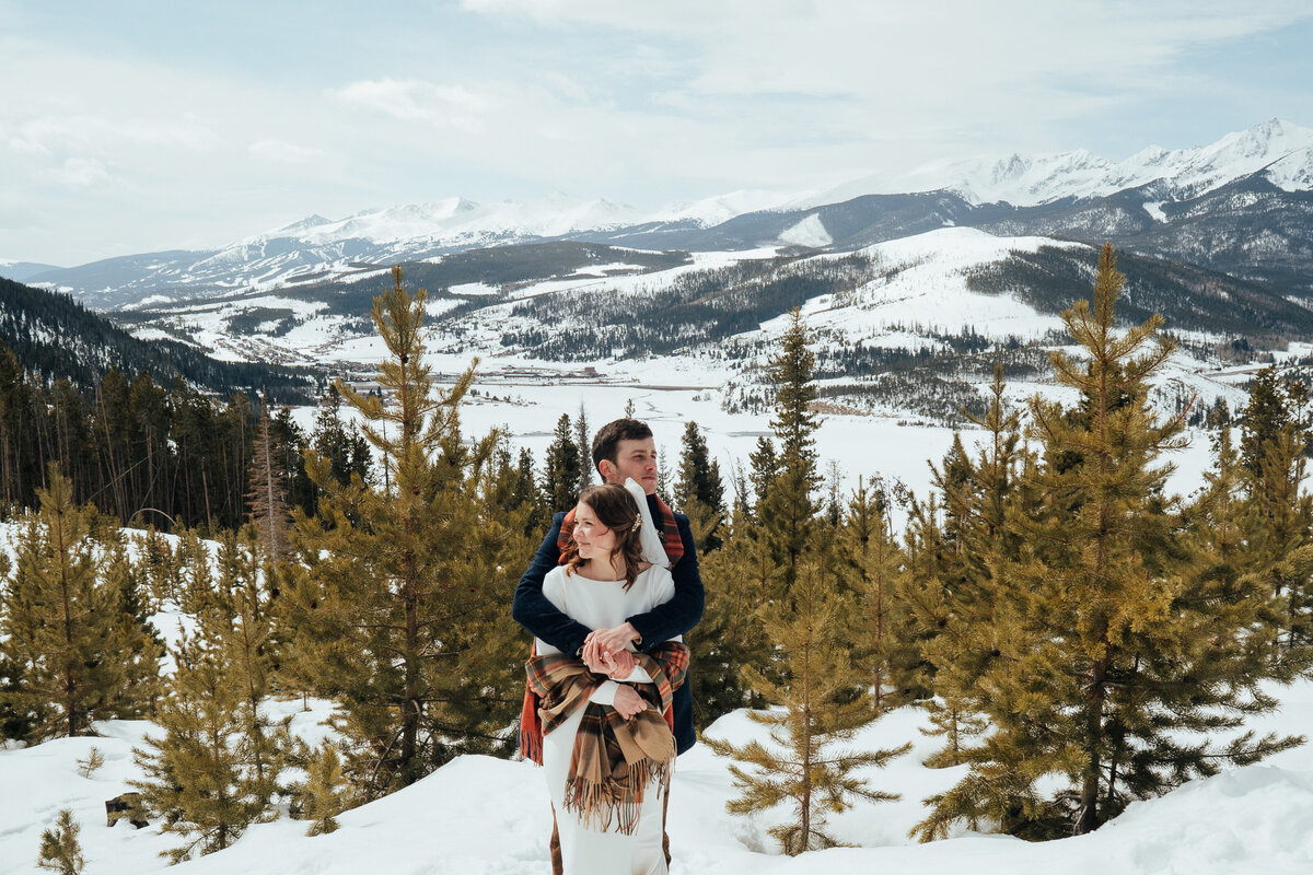 Couple cuddles in snowy forest with sweeping mountain views at Sapphire Point elopement.
