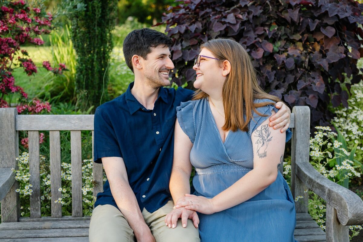 engaged couple sit on bench smiling at each other 