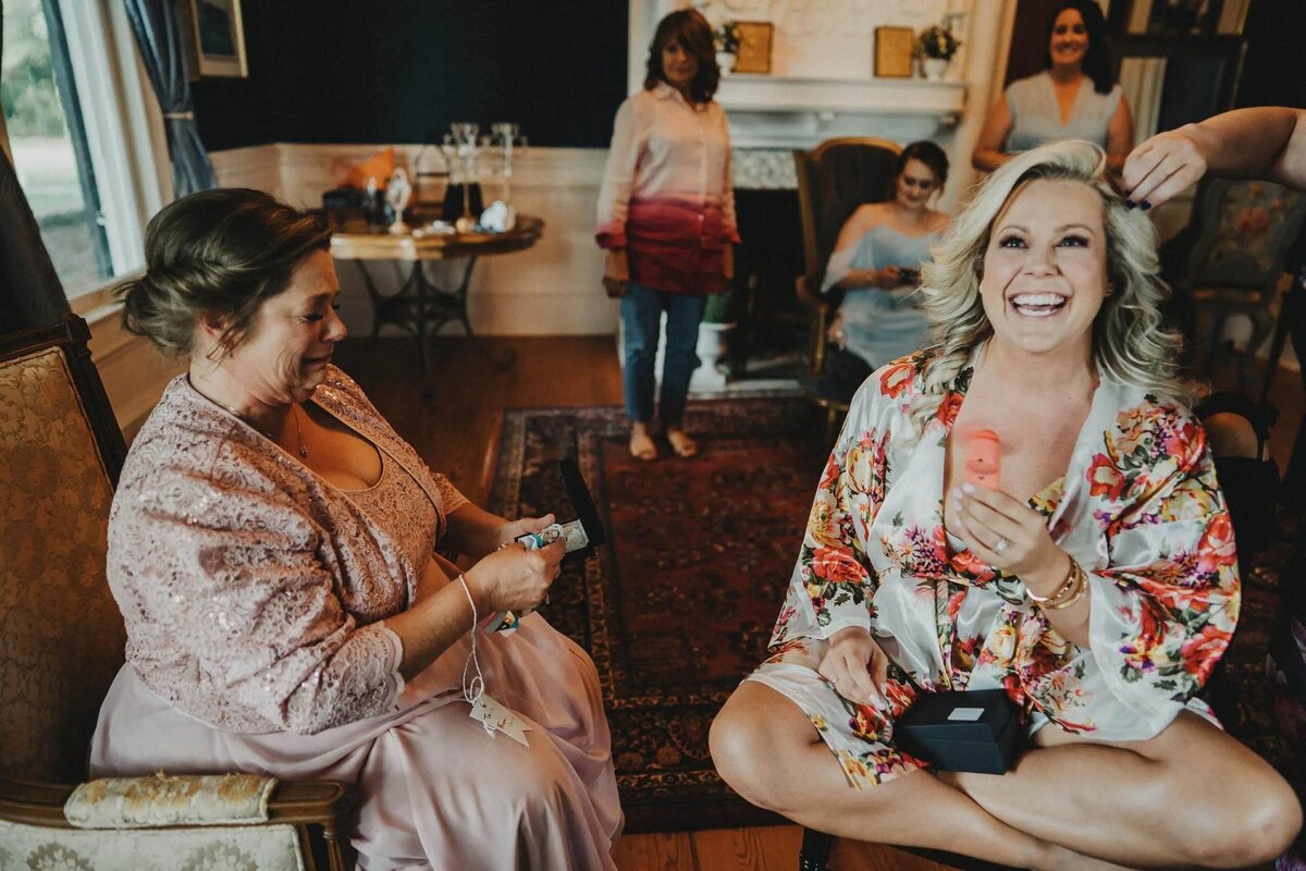 A bride, sitting in a robe, laughs heartily while holding a floral bouquet, with a woman sitting beside her and others in the background