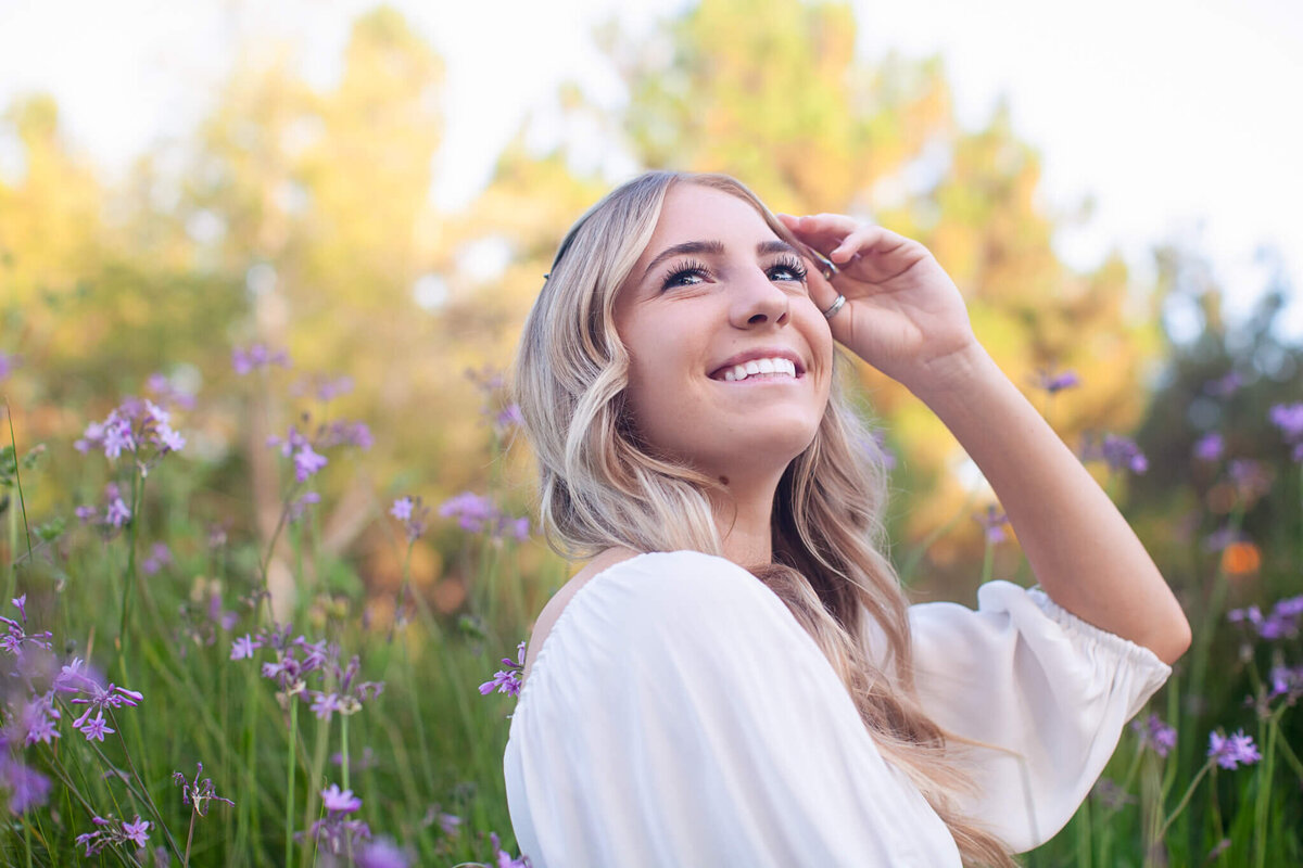 A woman with long blonde hair smiles while sitting in a field of purple flowers. She wears a white top and gestures with her hand, surrounded by greenery and blurred trees in the background. The sky is clear and the lighting is soft and natural.