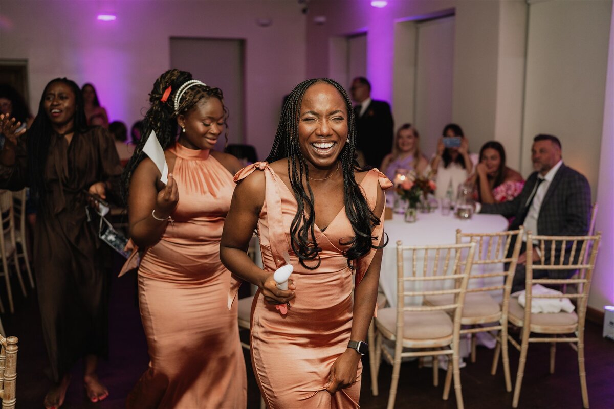 A large group of wedding guests dance in a line and walk through a large venue space. Fairy lights hang around them.
