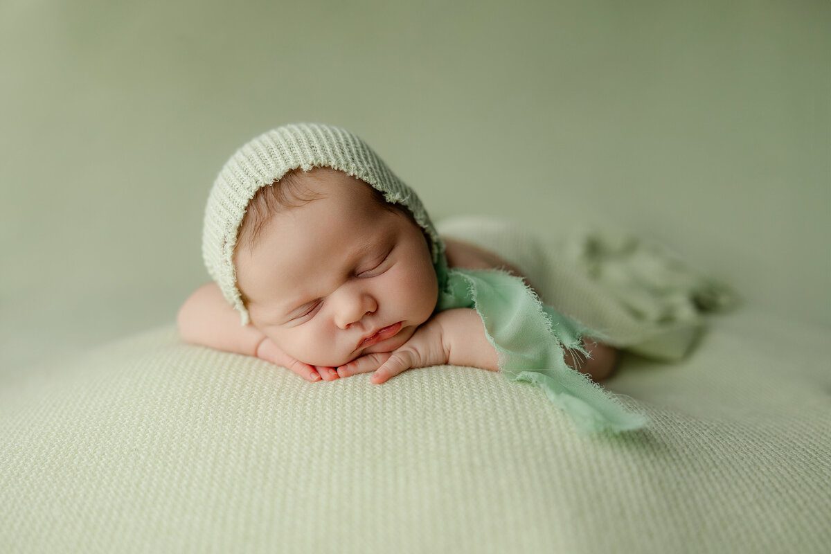 Newborn baby sleeping on a light green knit backdrop, styled with a cream bonnet and soft green wrap. Captured by Inland Empire newborn photographer Kaitlyn Dawn Photography.