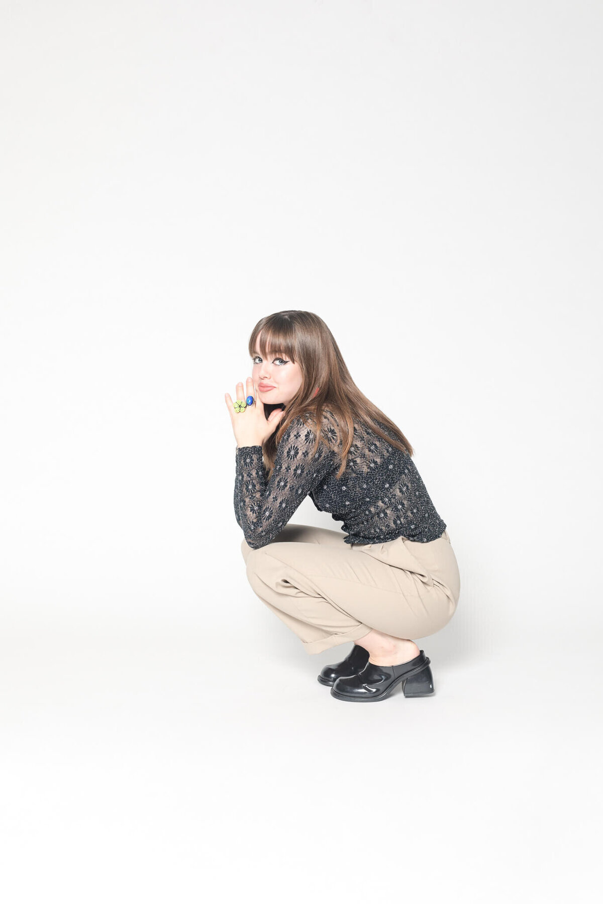 A woman with long brown hair crouches against a white background. She is wearing a black lace top, beige pants, and black shoes. She is holding a small green object in her hand near her face.
