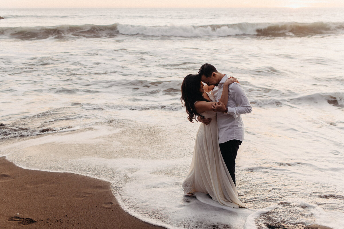 A couple embraces and kisses on a California beach at sunset during a photoshoot, with waves gently approaching their feet and the sun setting over the horizon, creating a warm, romantic atmosphere.