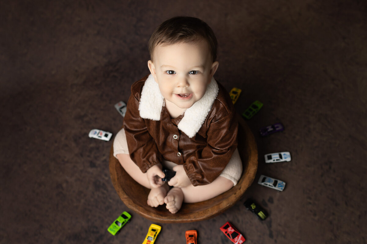 Toddler boy in a faux leather aviator jacket sitting in a wooden bowl, smiling up at the camera and surrounded by colorful toy cars during his Dallas milestone photographer session.