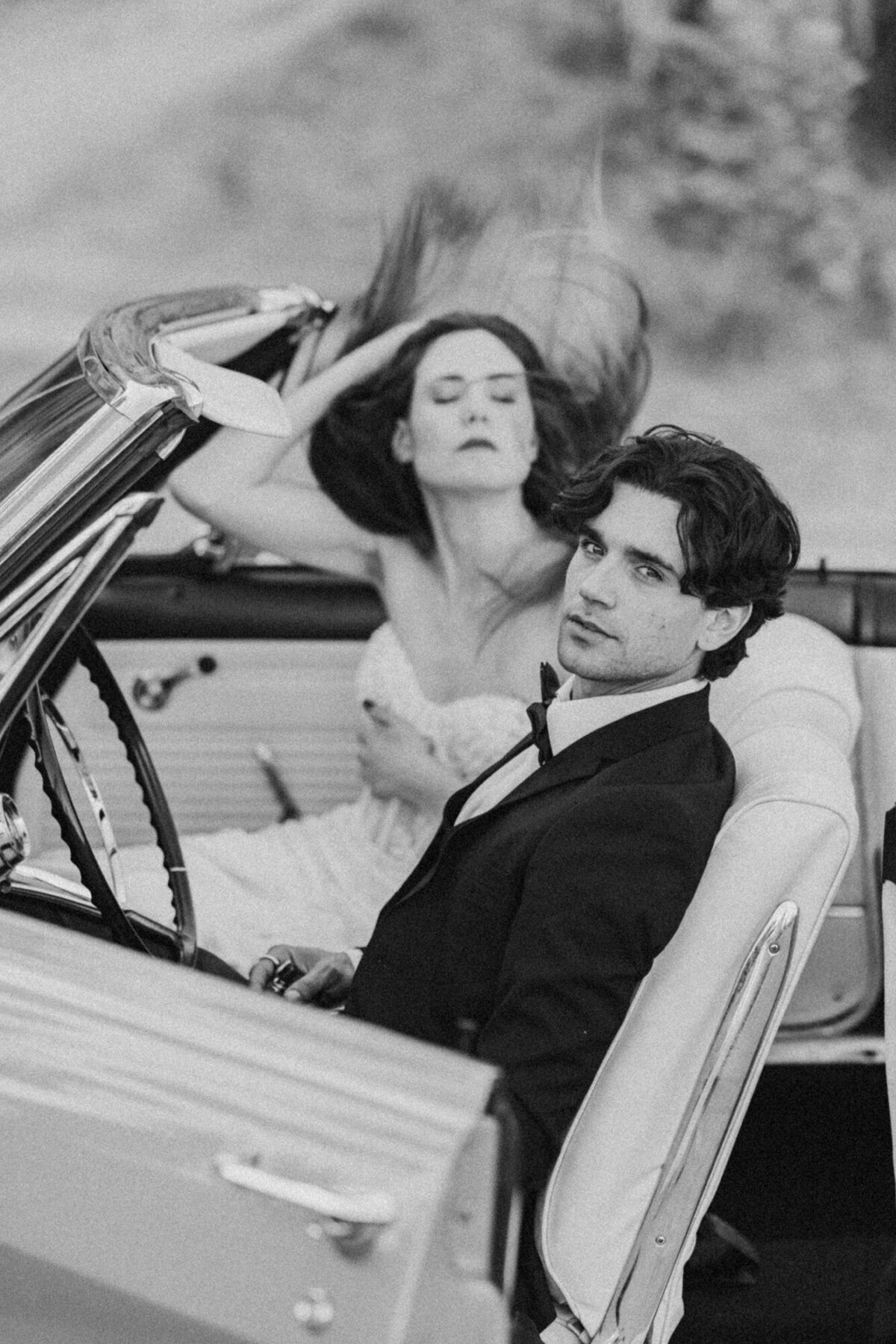 Black and white image of couple relaxing in a car, hand-in-hand, evoking a sense of calm connection and retro cool.
