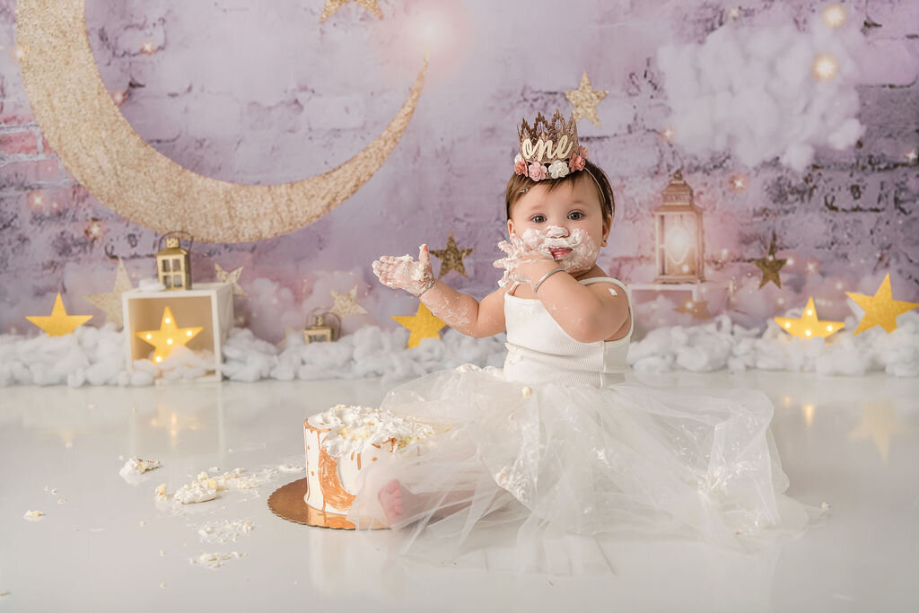 Portrait Photographers CT document joyful cake smash moment with baby girl covered in frosting wearing ivory tulle dress and floral crown against lavender backdrop with golden moon and stars