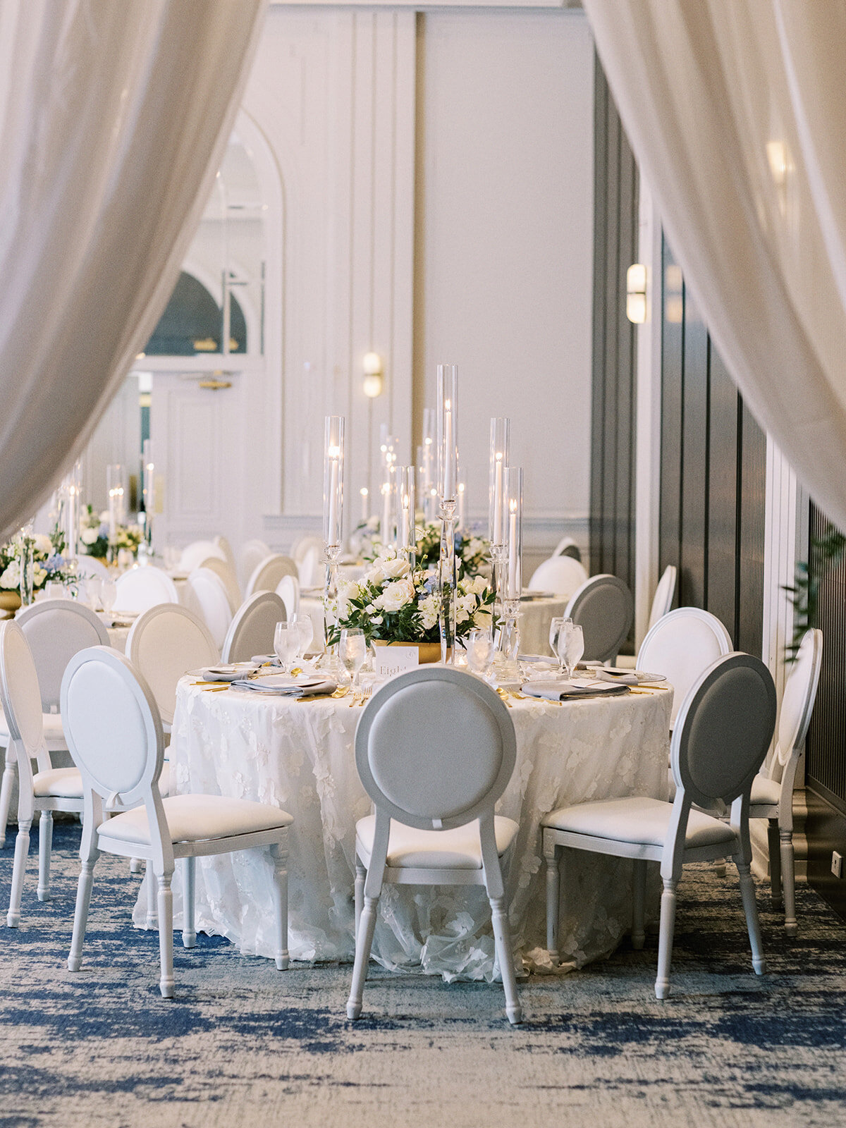 Elegant dining table setup with white chairs, floral centerpiece, and tall candleholders, framed by draped curtains in a sophisticated room with blue and white decor—perfect for a Banff Alberta wedding or any destination wedding in Canada.