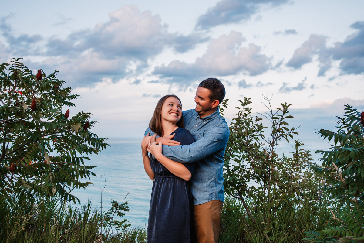 man holds woman from behind while they laugh on the bluffs of lake michigan