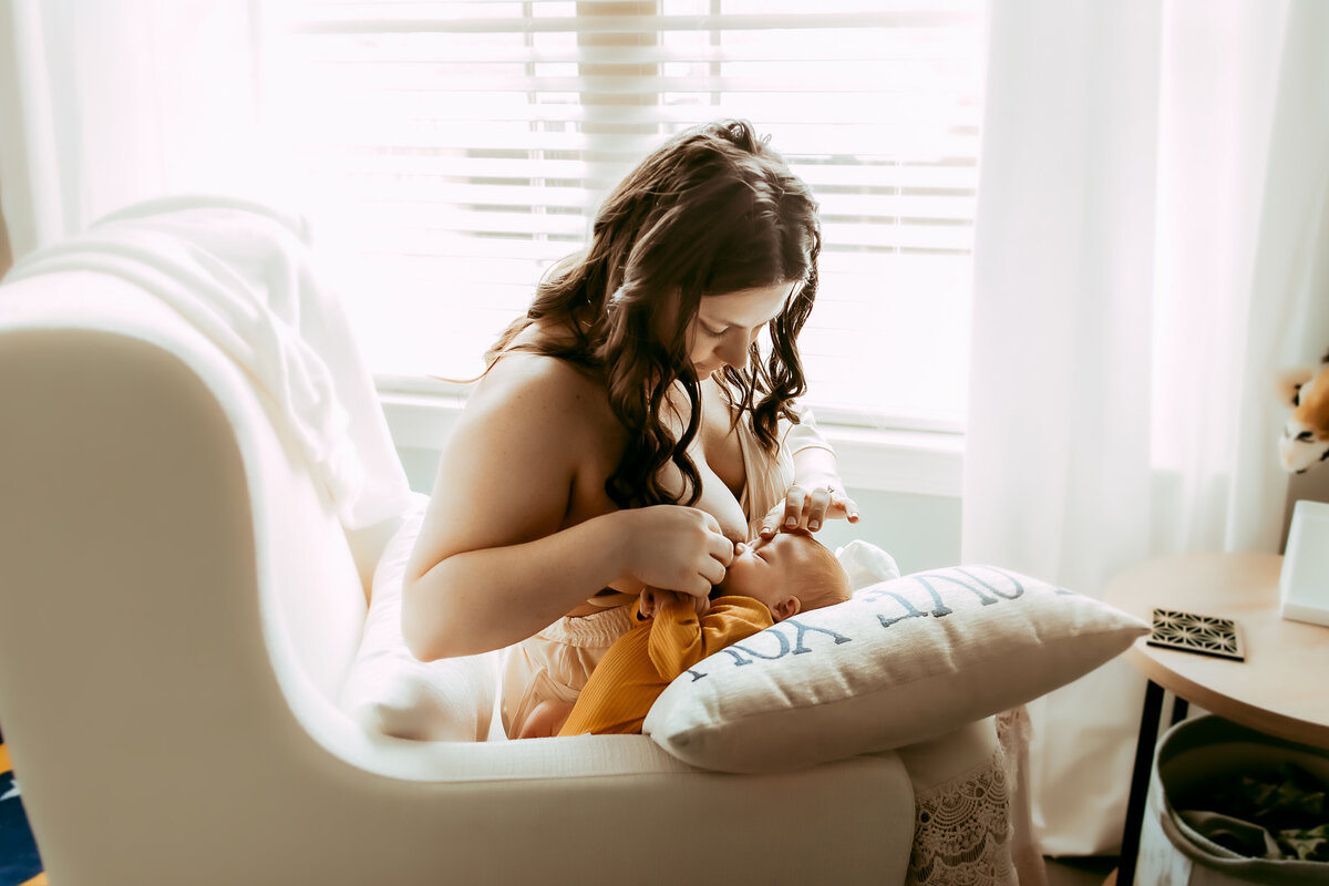 A woman breastfeeds a baby in a softly lit room, sitting on a white chair near a window with blinds, holding a pillow with text.
