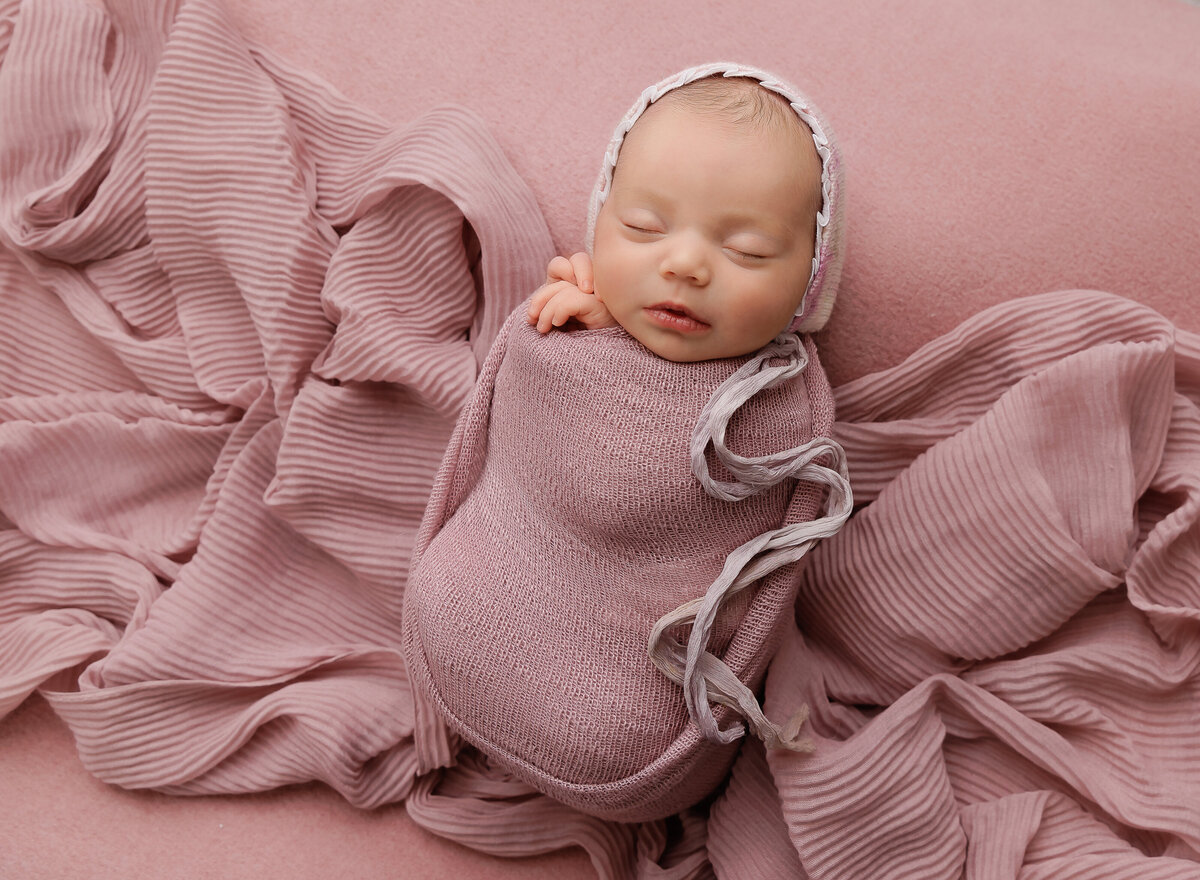 Brooklyn newborn photo captures baby girl swaddled in pink with her hands peeking out of the swaddle. She is wearing a knit cap. Captured by premier Brooklyn NY newborn photographer Chaya Bornstein Photography.