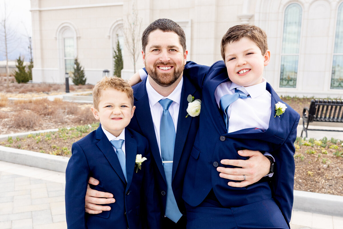 Groom smiles while standing between his two young sons, one of whom is playfully lifted in his arms, all wearing matching blue suits and boutonnieres.