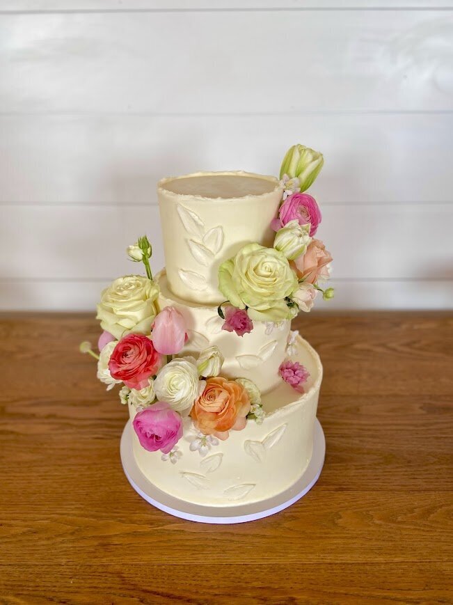 A three tier cake sits on a wooden farm table. The frosting is all white with rough edges and pallet knife leaf accents. Two clusters of spring florals also decorate the cake. 