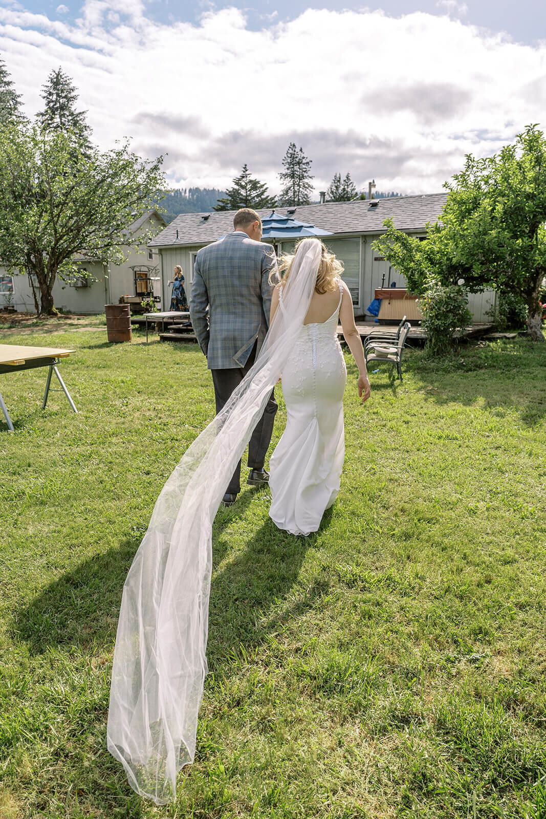A bride and groom walk towards a house with the bride's veil blowing in the wind.