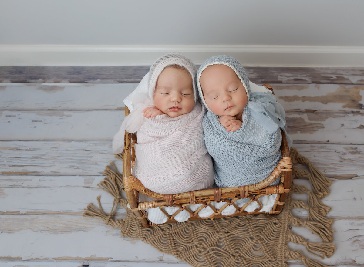Twin brother and sister are wrapped in blue and pink swaddles with their hands peeking out of the swaddle. They are placed together in a basket with their heads resting together. Captured by top Brooklyn newborn photographer Chaya Bornstein