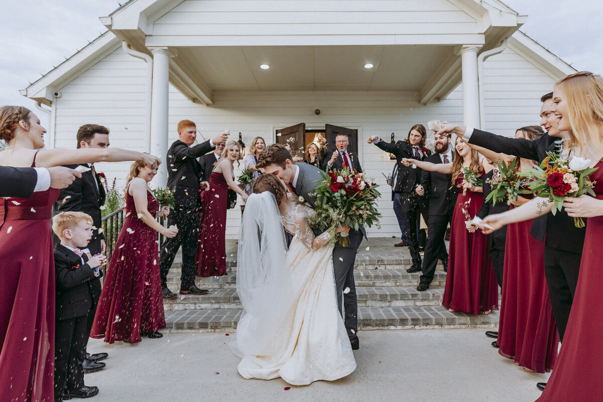 Groom dip kisses bride while they exit the chapel and wedding party throws flowers over them