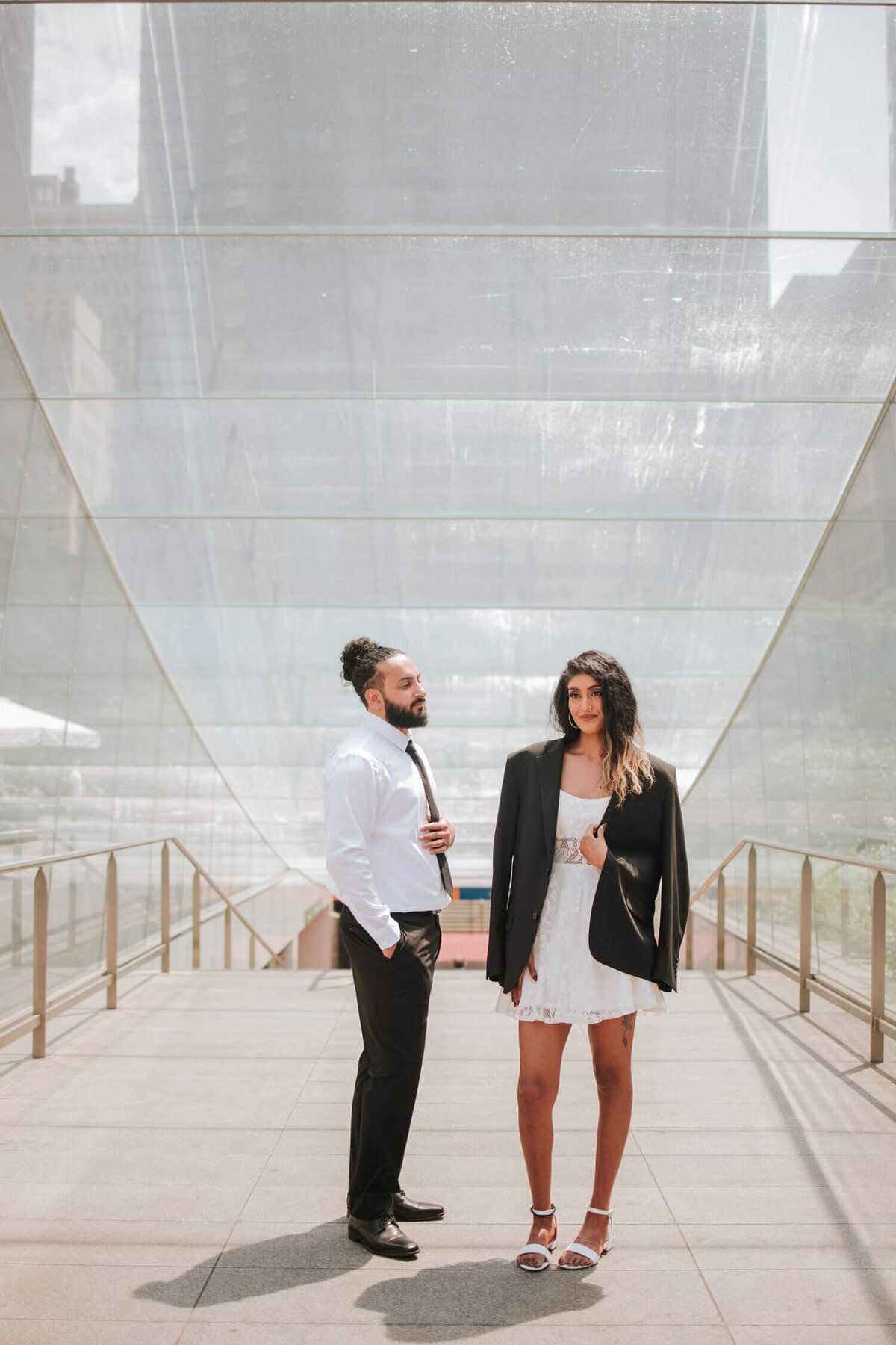 "A stylish couple poses at a modern glass structure in Philadelphia, exuding confidence and elegance in contemporary attire