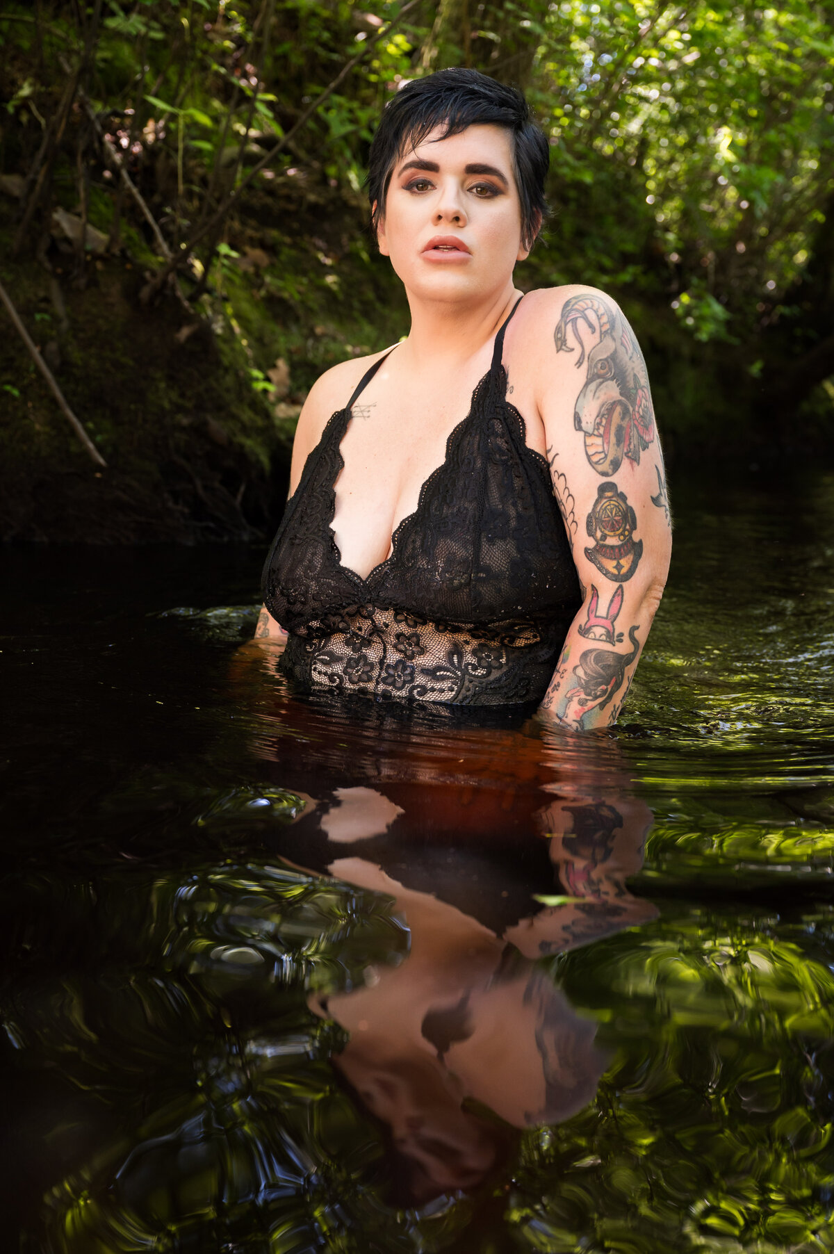 A person with short dark hair and tattoos is partially submerged in water, wearing a black lace top, with greenery in the background.