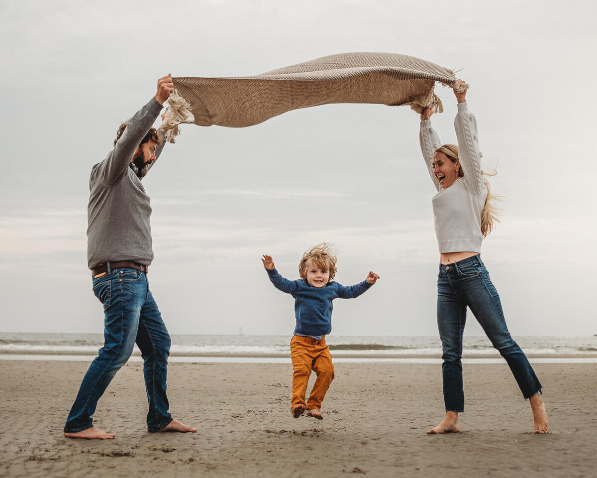 This Charleston family photoshoot on beach in Sullivan's Island shows a mother and father lifting a blanket over their son while he jumps for joy.