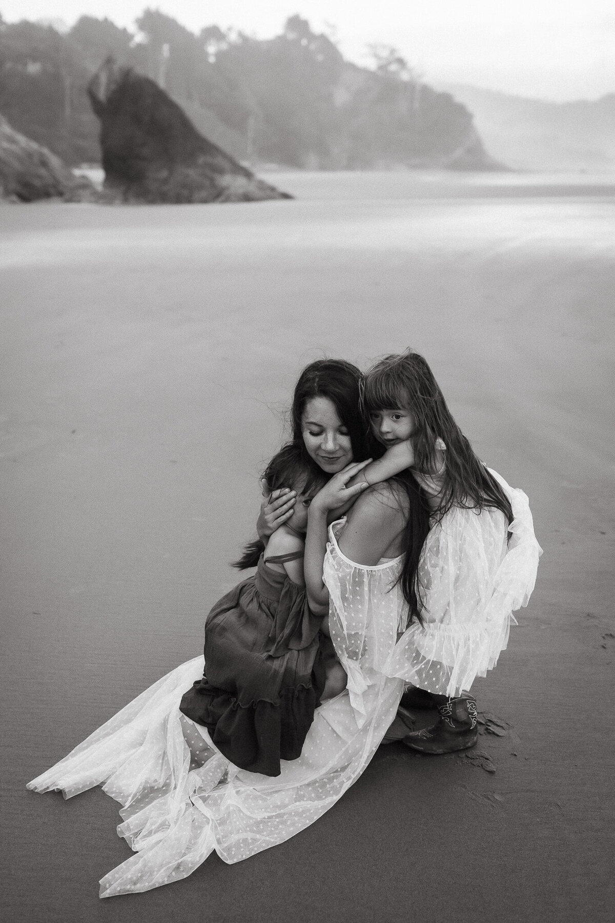Motherhood portrait on beach of mom embracing baby and other child hugs mom around shoulders